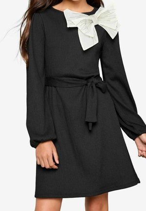 Sorte langærmede kjole med struktureret stof, indsnævret talje og en stor hvid sløjfeaccent med metalliske tråde ved halsudskæringen.