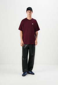 Jauns vīrietis valkā melnu cepuri, bordo Adidas t-kreklu, melnus Adidas treniņbikses un zilos Adidas kedus, stāvot pret vienkāršu baltu sienu.