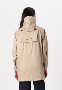 Beżowa wodoodporna kurtka z wysokim kołnierzem i regulowanym kapturem, z logo z tyłu oraz długimi rękawami. Gładki, lekki materiał.