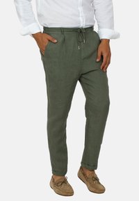 Pantaloni di lino verdi con vita regolabile, tasche laterali, design a gamba affusolata e tessuto leggero. Abbinati a scarpe beige senza lacci.