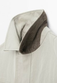 Bežový límec bundy vyhrnutý nahoru, odhalující měkkou hnědou semišovou vnitřní podšívku a detaily švů na povrchu látky.