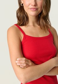 Rdeča majica brez rokavov s tankimi naramnicami, izdelana iz strukturirane tkanine z rahlim vzorcem. Modelka ima prečkane roke, kar prikazuje prileganje oblačila.