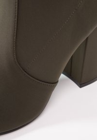 Gros plan d'une botte cheville vert olive foncé avec des coutures détaillées et un talon épais sur un fond blanc.