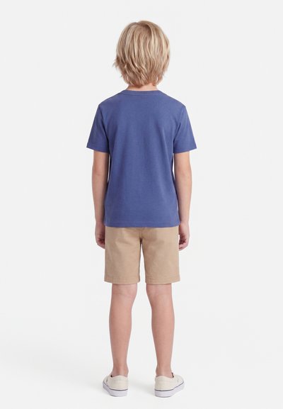 Ξανθό παιδί που στέκεται με την πλάτη, φοράει μπλε κοντομάνικη μπλούζα, μπεζ σορτς και λευκά αθλητικά σε απλό λευκό φόντο.