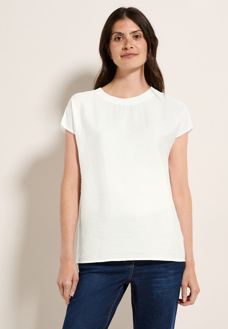 Chemise blanche à manches courtes avec un col rond, en tissu texturé et coupe ample, associée à un jean bleu foncé.