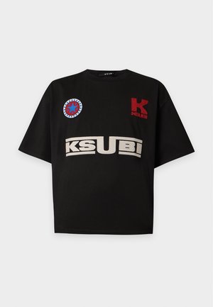 Homme portant un t-shirt noir à motif graphique, un jean large sombre avec des revers, des lunettes de soleil noires et des baskets noires et blanches, debout devant un fond uni.