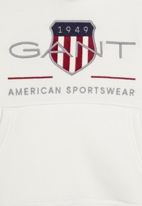 White hoodie with embroidered blue and red shield logo. Features "GANT" text and "AMERICAN SPORTSWEAR" below. Soft texture, kangaroo pocket.