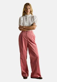 Frau steht mit verschränkten Armen, trägt eine weiße, rüschenbesetzte Bluse mit kurzen Ärmeln und eine rosa, weit geschnittene Hose vor einem schlichten Hintergrund.