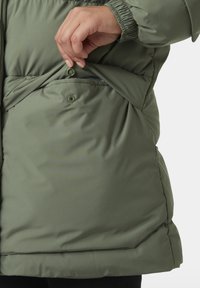 Chaqueta de plumas de color verde oliva con un cierre frontal de cremallera y detalles de bolsillos ocultos. La tela es suave, con una textura sutil y puños elásticos.