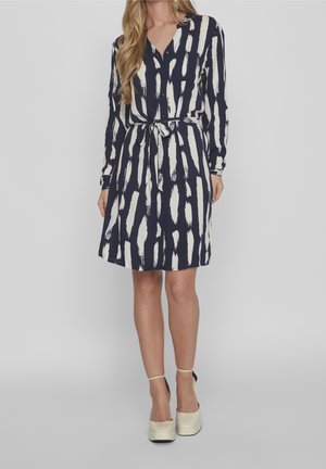 Kvinde iført en mørkeblå kjole med hvide lodrette penselstrøgsmønstre og et bælte i taljen, kombineret med cremefarvede plateauhæle med ankelstropper.