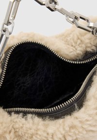 Taška z umelého kožušiny s jemnou krémovou textúrou a čiernym koženým vnútrom, vybavená strieborným zipsom a masívnou striebornou reťazou ako rúčkou.