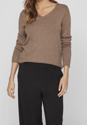 Femme portant un pull marron clair à manches longues avec un col en V et un pantalon noir, debout devant un fond uni et clair.