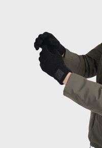 Svart stickade handskar med textilstruktur och en fyrkantig logotyp på handleden, som bärs av en person som justerar sitt grepp.