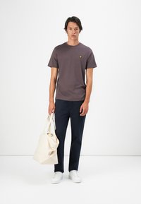 Mann in braunem T-Shirt, dunkler Hose und weißen Turnschuhen, hält eine beige Tote-Bag und steht vor einem schlichten weißen Hintergrund.