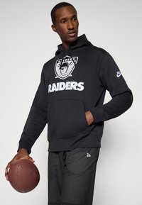 Čierna mikina s kapucňou s logom Raiders a bielym textom, s vreckom na klokan a sťahovacou kapucňou. Nosené s čiernymi sieťovanými šortkami, drží futbal.