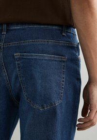 Jeans de denim azul oscuro con costuras en contraste, un bolsillo trasero y una cinturilla. La tela es suave con una ligera textura.