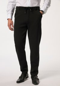 Zwarte op maat gemaakte broeken met een trekkoord in de taille, taps toelopende pijpen en twee zijzakken, gecombineerd met zwarte leren schoenen en een witte shirt.