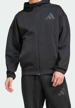 Homme portant une veste à capuche noire zippée et un pantalon assorti avec des logos Adidas discrets sur la poitrine et la cuisse.