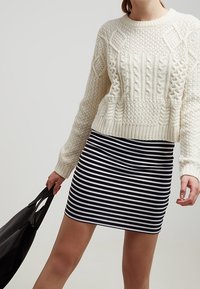 Kermaankeltainen palmikkoneulepusero, jossa on kohokuvioituja kuvioita, yhdistetty tiukkaan mustavalkoraidalliseen minihameeseen. Malli pitää kädessään mustaa laukkua.