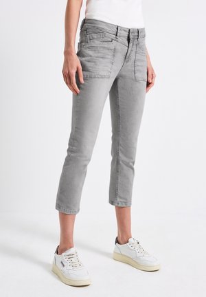 Person iført kortgrå jeans med forlommer og hvide sneakers, stående mod en ensfarvet hvid baggrund.