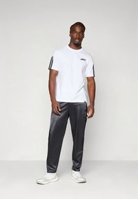 Valge lühikeste varrukatega t-särk musta logoga, hallide satiinist joggerite ja valgete tossudega, neutraalse tausta ees seismas.