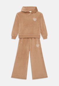 Sweatshirt à capuche en velours beige avec un logo sur la poitrine, associé à un pantalon large avec des poches latérales et une taille avec cordon de serrage.
