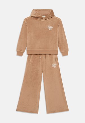 Tan velour hooded sweatshirt with a logo on the chest, paired with wide-legged pants featuring side pockets and a drawstring waistband.
