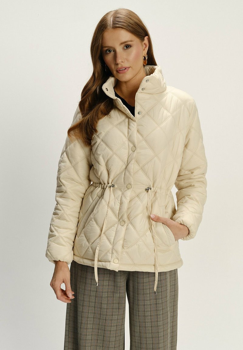 Frau in einer beigen gesteppte Jacke mit Kordelzug in der Taille, die mit einer Hand in der Tasche steht, kombiniert mit grauen, karierten weitgeschnittenen Hosen.