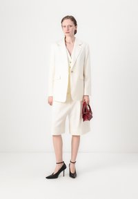 Elfenbeinfarbener, taillierter Blazer mit passenden Shorts und strukturierter Passform. Schwarze High-Heels und eine kleine rote Handtasche runden das Outfit ab.