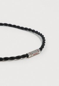 Sort tvistet læderarmbånd med en sølvspænde med røde accenter og "HUGO" indgraveret. Enkel og elegant design.