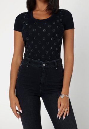 Femme portant un haut noir à manches courtes avec de petits motifs circulaires et un jean noir taille haute, montrant un bracelet et une bague à la main droite.