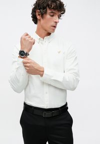 Biela košeľa s dlhými rukávmi a malým zlatým emblémom. Doplnok čiernymi spoločenskými nohavicami a striebornými hodinkami.