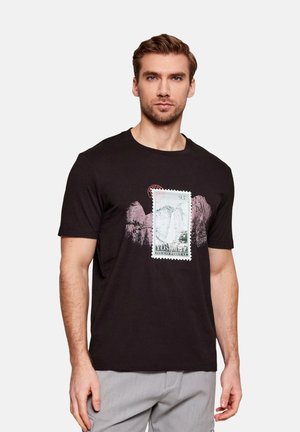 Hombre con camiseta negra con estampado de Yosemite y gráfico de montaña, posando contra un fondo liso y claro.