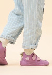 Lila, weiche Schuhe mit zwei Klettverschlüssen und runder Zehenpartie. Strukturierter Stoff mit einem Logo-Detail an der Ferse. Getragen mit hochgekrempelten hellen Jeans.