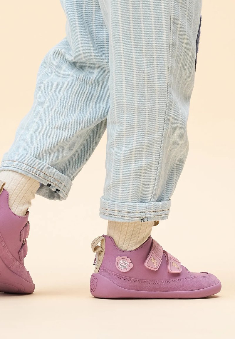 Lila, weiche Schuhe mit zwei Klettverschlüssen und runder Zehenpartie. Strukturierter Stoff mit einem Logo-Detail an der Ferse. Getragen mit hochgekrempelten hellen Jeans.