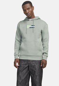 Haut en coton vert clair avec poche frontale, cordons de serrage et impression de logo blanche. Associé à un pantalon en denim foncé.