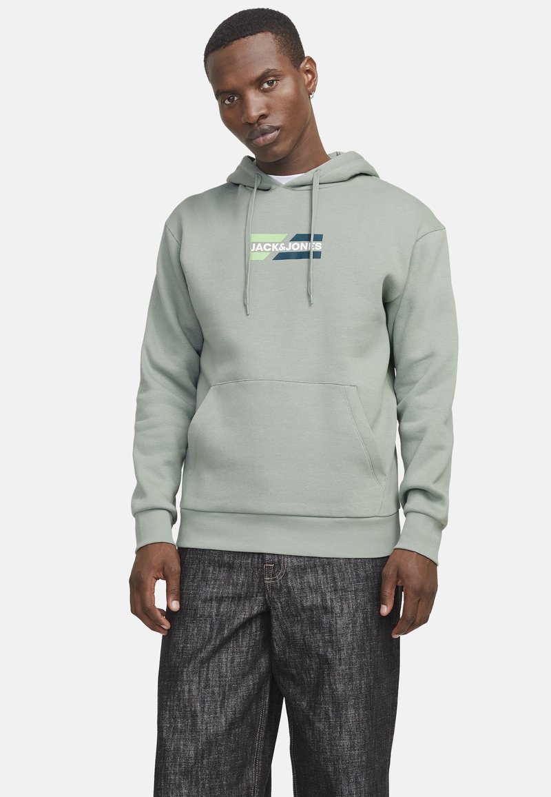 Haut en coton vert clair avec poche frontale, cordons de serrage et impression de logo blanche. Associé à un pantalon en denim foncé.