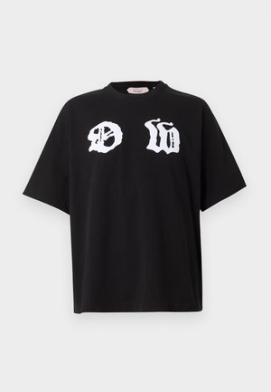 Mand stående med den ene hånd i lommen iført sort oversized T-shirt med hvidt grafisk tekst, sorte vidde bukser og sort-hvide sneakers.