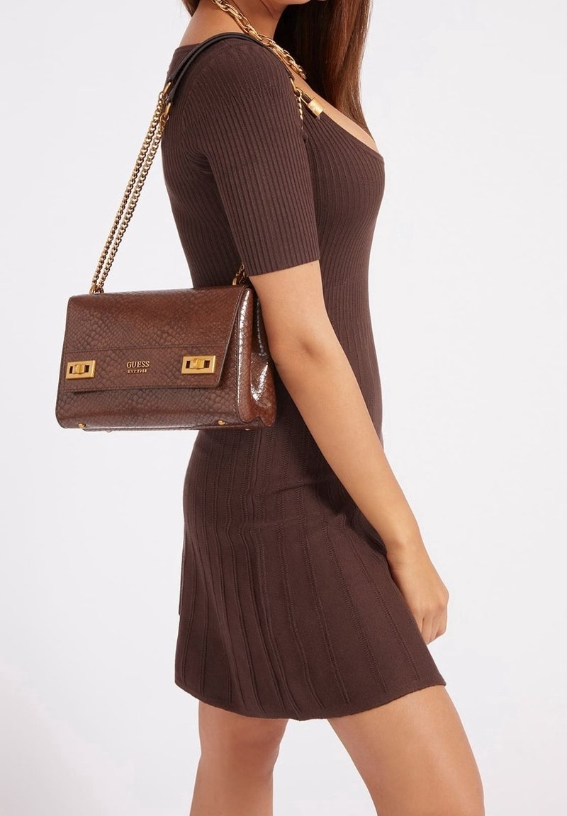 Donna in un vestito marrone a coste aderente che porta una borsa Guess in pelle marrone testurizzata con manico in catena dorata sulla spalla.