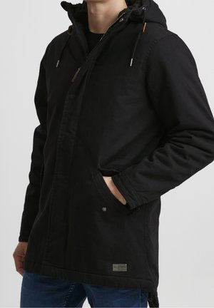 Homme portant une veste à capuche noire avec fermeture éclair et boutons, main dans la poche, associée à un jean bleu sur un fond uni.