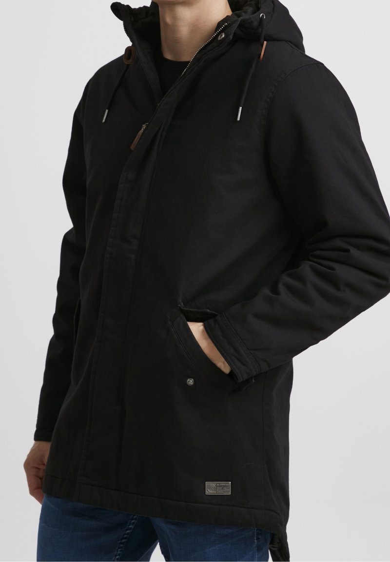 Homme portant une veste à capuche noire avec fermeture éclair et boutons, main dans la poche, associée à un jean bleu sur un fond uni.