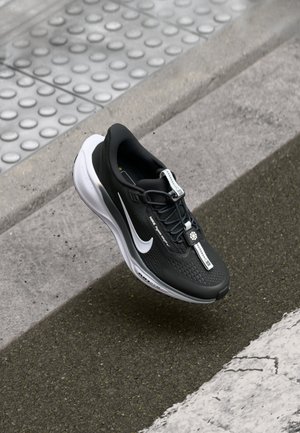 Scarpa da corsa Nike bianca e nera con swoosh bianco, appoggiata sul marciapiede vicino al bordo e all'indicatore tattile di superficie.