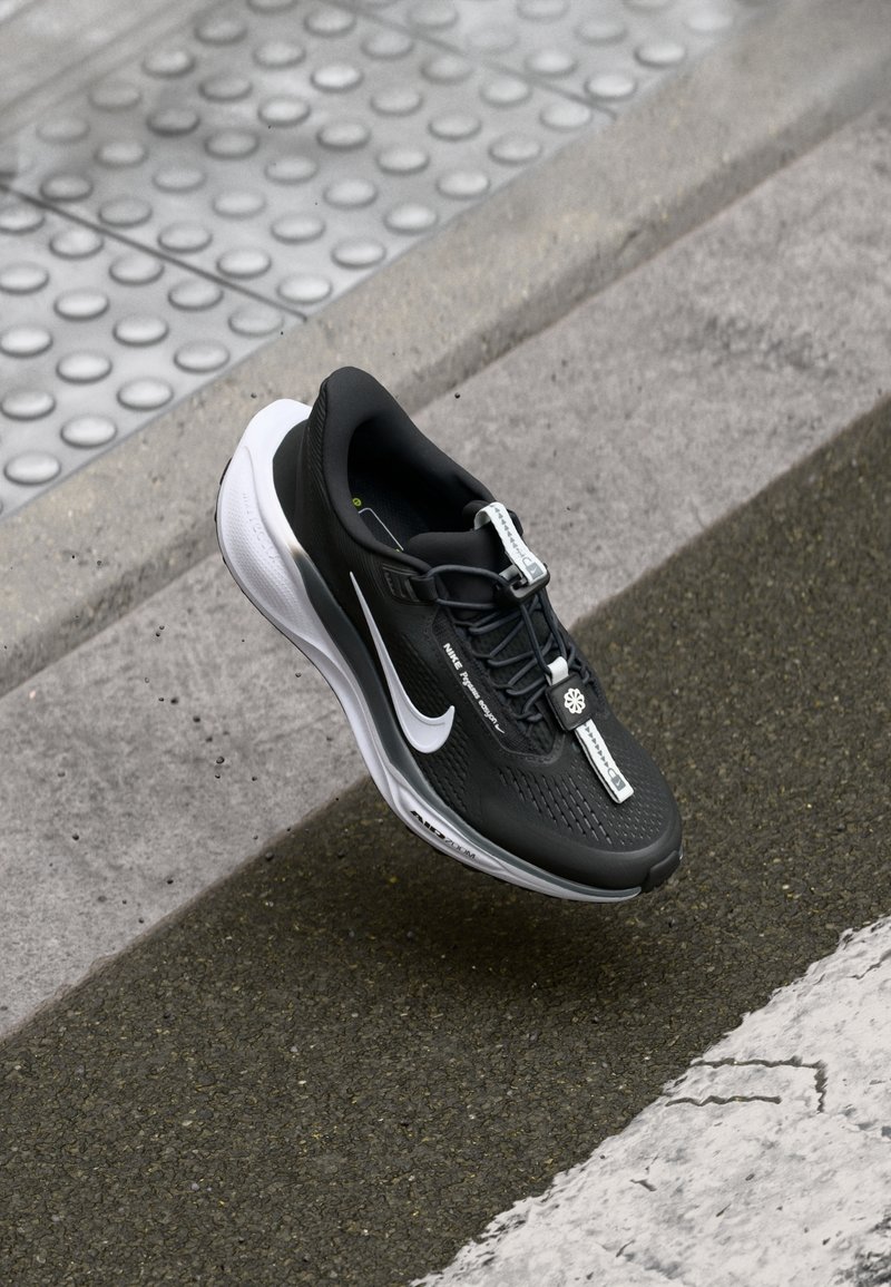 Chaussure de course Nike noire et blanche avec un swoosh blanc, reposant sur le trottoir près du bord et de l'indicateur tactile de surface.