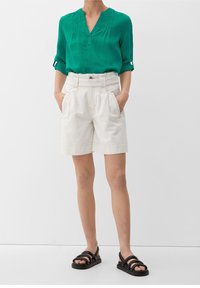 Grüne kurzärmlige Bluse mit V-Ausschnitt, kombiniert mit beigen hochgeschnittenen Shorts mit Falten und Seitentaschen, getragen mit schwarzen Sandalen.