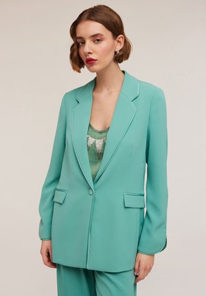 Turquoise blazer in smooth fabric with a fitted design, notch lapels, two front pockets, and a single button closure, styled over a green top.