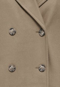 Manteau beige croisé à l'avant montrant quatre boutons marbrés et des détails de couture près du col et des boutonnières.
