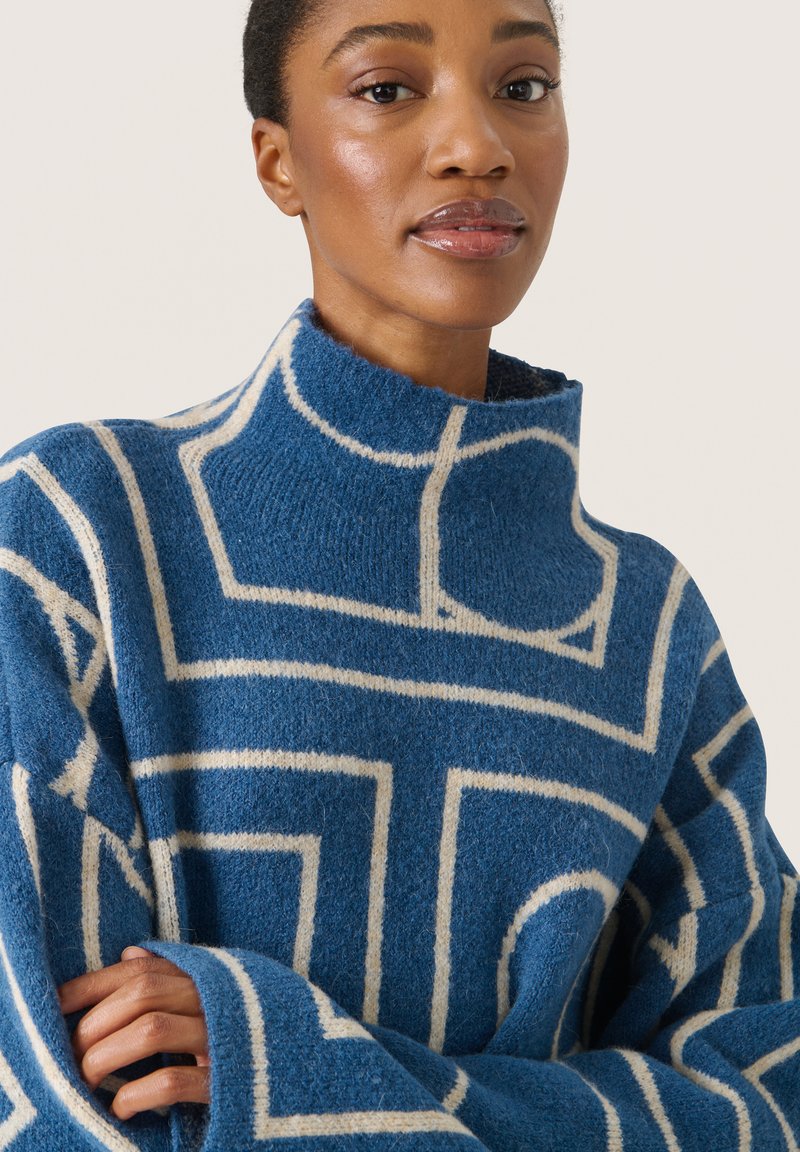 Blauer Wollpullover mit hohem Kragen, der ein weißes geometrisches Muster und lockere, weite Ärmel hat. Strukturierte Strickware.