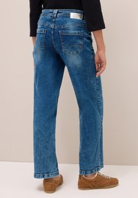Personne portant un jean droit bleu et des baskets brunes, debout et vu de dos contre un fond neutre.