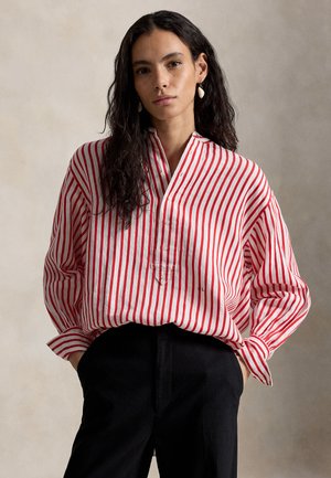 Ženska z dolgimi temnimi lasmi nosi ohlapno rdeče-belo črtasto bluzo in črne hlače, stoji z rokami v žepih na nevtralni podlagi.