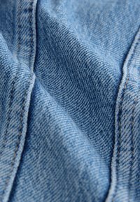 Detailný záber svetlomodrej rifľoviny zobrazujúci detailné tkanie a dvojito prešité švy s oranžovou niťou.